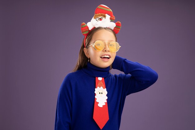 Little girl in blue turtleneck with red tie and  funny christmas rim on head looking at camera with happy face smiling cheerfully standing over purple background