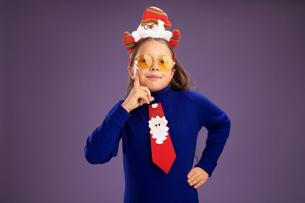 Little girl in blue turtleneck with red tie and  funny christmas rim on head looking at camera with smile on face standing over purple background