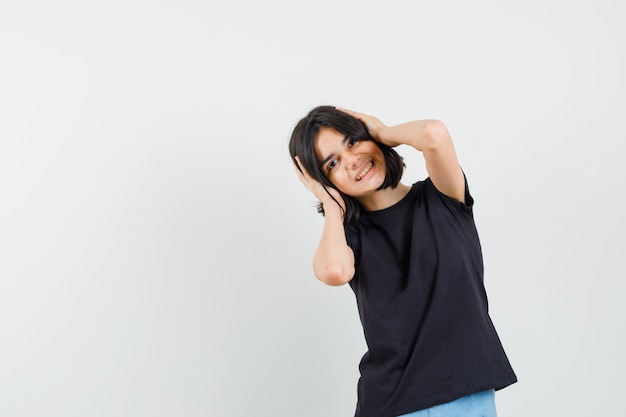 Little girl clasping head with hands in black t-shirt, shorts and looking cheerful , front view.