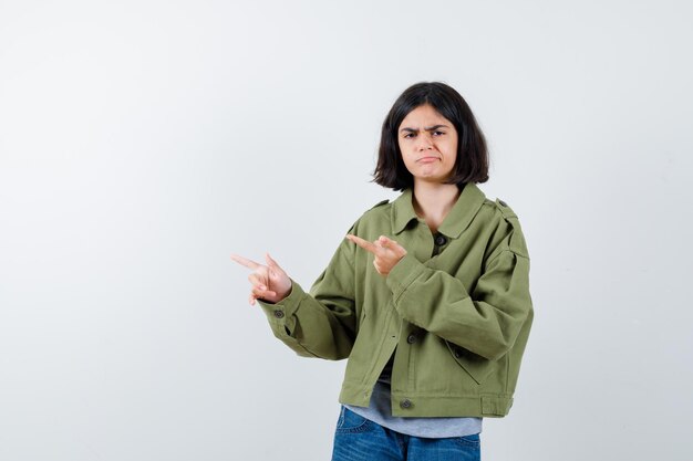 Little girl in coat, t-shirt, jeans pointing to the left side and looking pensive , front view.