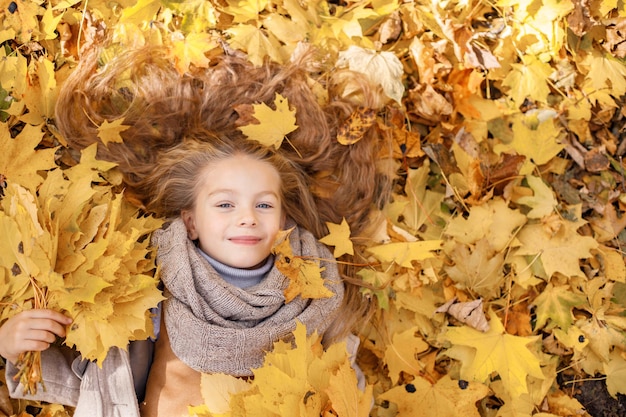 Little girl in fashion clothes in autumn forest. Girl lying in a yellow leaves. Girl wearing brown scarf and looking in camera.