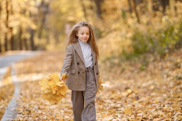 Little girl in fashion clothes walking in autumn park. Girl holding a yellow leaves. Girl wearing brown costume with jacket.