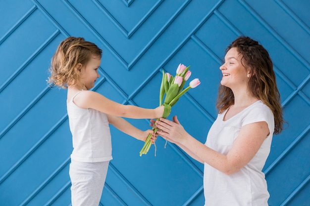 Free photo little girl giving pink tulips flowers to her mom on mother's day in front of blue background