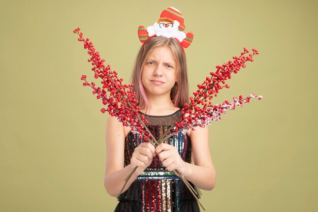 Little girl in glitter party dress and headband with santa holding branches with red berries looking at camera making wry mouth with disappointed expression standing over green background