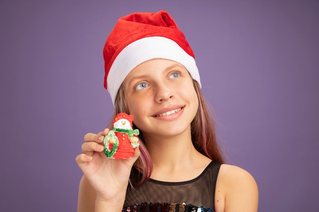 Little girl in glitter party dress and santa hat showing christmas toy looking up with smile on face standing over purple background