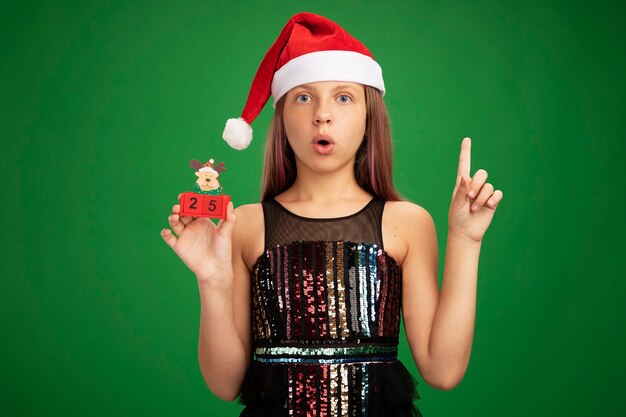 Little girl in glitter party dress and santa hat showing toy cubes with date twenty five looking surprised showing index finger standing over green background