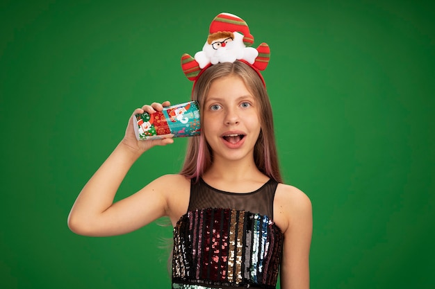 Little girl in glitter party dress and santa headband holding colorful paper cup over her ear smiling cheerfully standing over green background