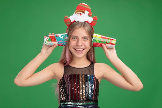 Little girl in glitter party dress and santa headband holding colorful paper cups over her ears smiling cheerfully standing over green wall