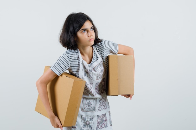 Little girl holding cardboard boxes in t-shirt, apron and looking hesitant ,