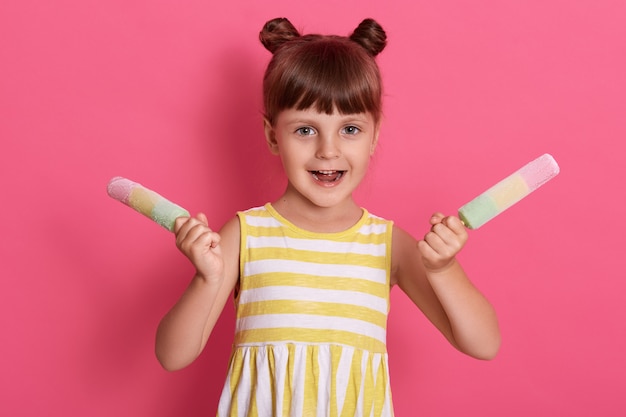 Free photo little girl holding colorful ice lollies, having excited facial expression, posing with happy look, wearing white and yellow stripped summer dress against pink wall.