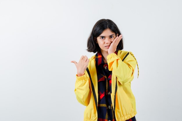 Little girl holding hand on cheek while pointing to the left side with thumb in checked shirt, jacket and looking confident