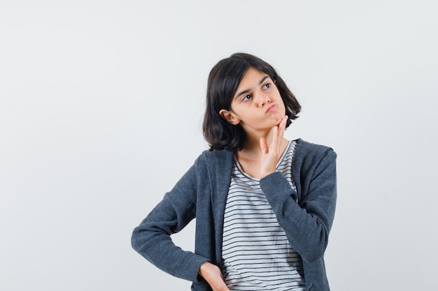 Little girl holding hand on chin in t-shirt, jacket and looking hesitant