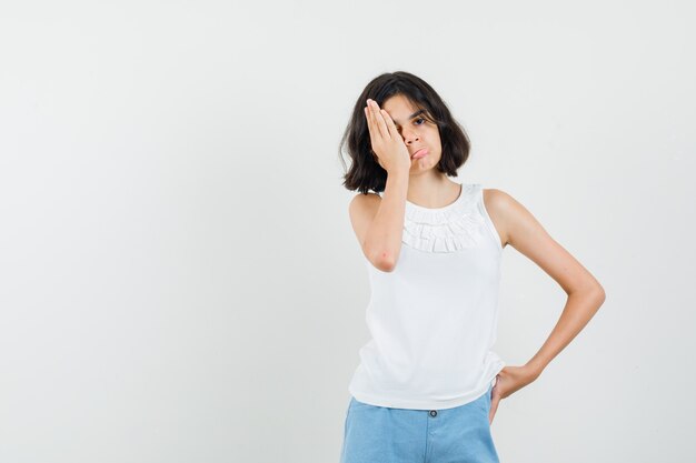 Little girl holding hand on eye in white blouse, shorts and looking resentful. front view.