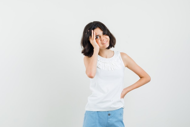 Little girl holding hand to face in white blouse, shorts and looking cheerful , front view.