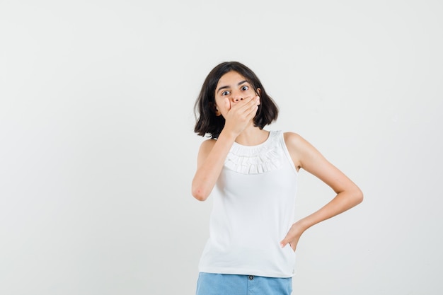 Little girl holding hand on mouth in white blouse, shorts and looking surprised , front view.