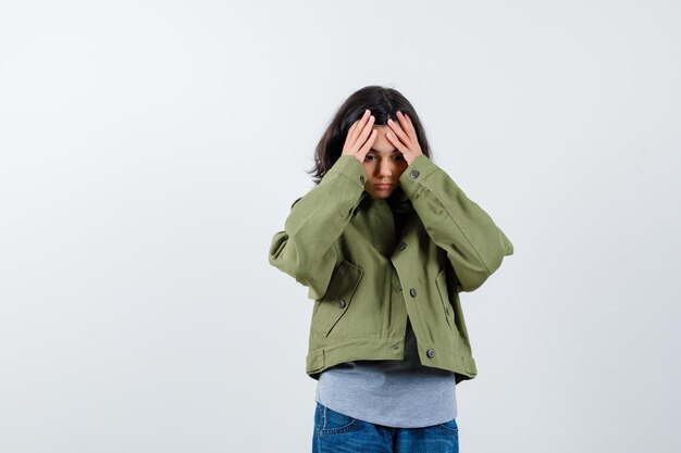 Little girl holding hands on forehead in coat, t-shirt, jeans and looking forgetful , front view.