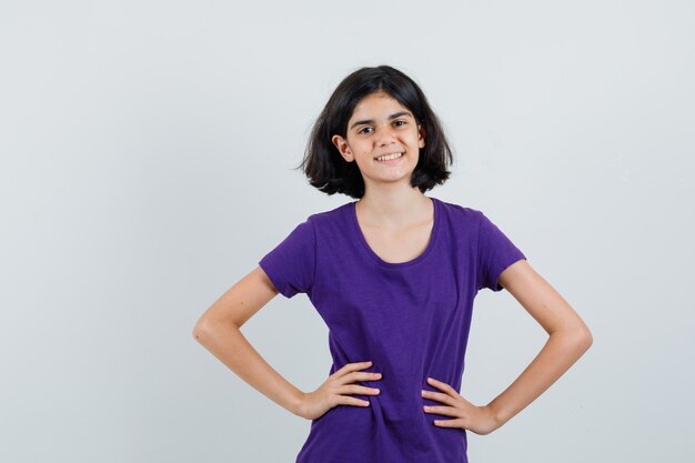 Little girl holding hands on waist in t-shirt and looking joyful ,