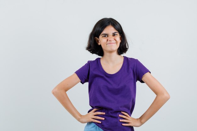 Little girl holding hands on waist in t-shirt, skirt and looking glad ,