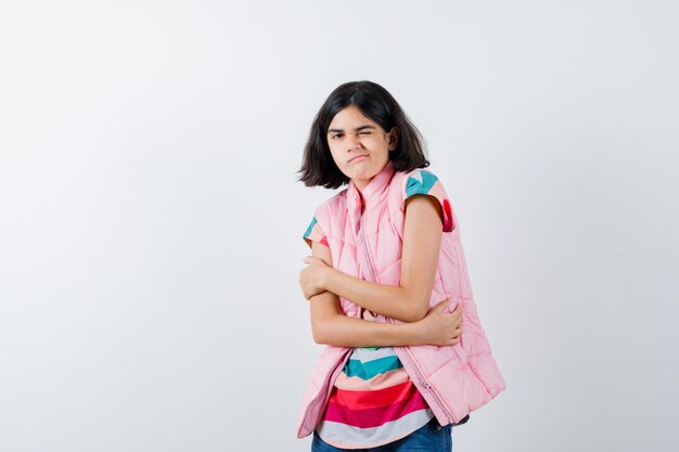 Little girl hugging herself in t-shirt, puffer vest, jeans and looking puzzled , front view.