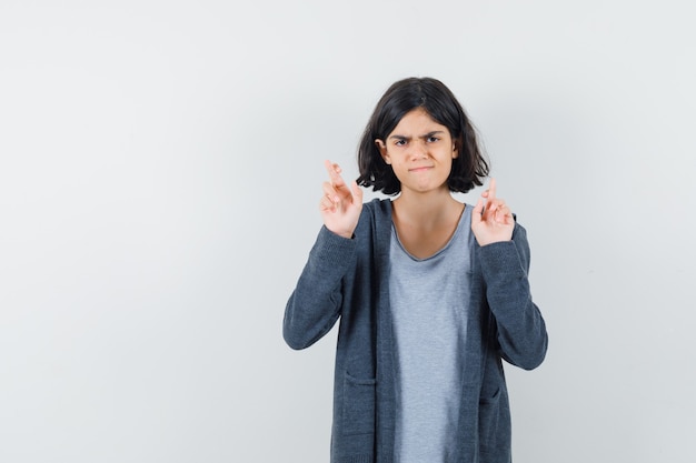 Little girl keeping fingers crossed in t-shirt, jacket and looking doubtful.