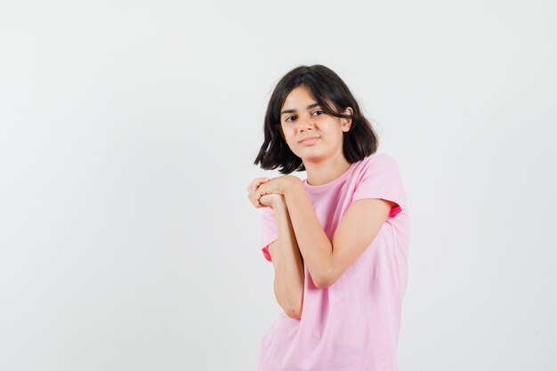 Little girl keeping hands clasped in pink t-shirt and looking cute , front view.