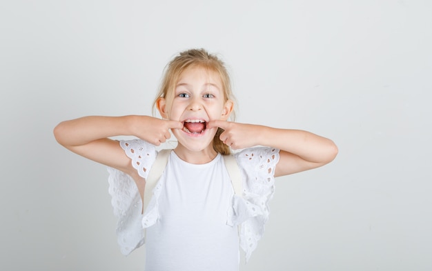 Little girl opening mouth with fingers in white t-shirt and looking funny.