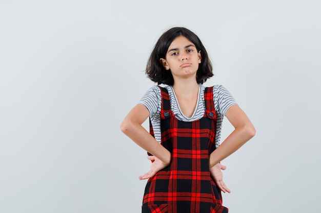 Free photo little girl in pinafore dress holding hands on waist and looking sad ,