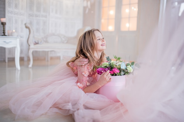 Little girl in pink dress holds box with roses sitting on the floor in a luxury room