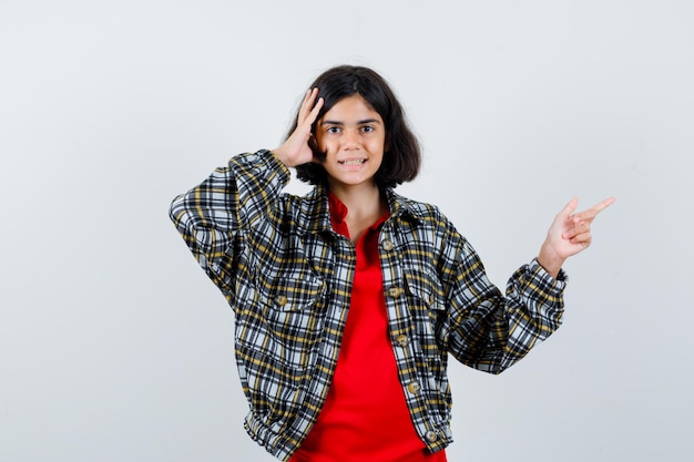 Free photo little girl pointing aside while holding hand on head in shirt,jacket and looking talkative , front view.