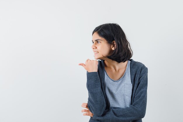 Little girl pointing aside with thumb in t-shirt, jacket and looking dreamy. front view.