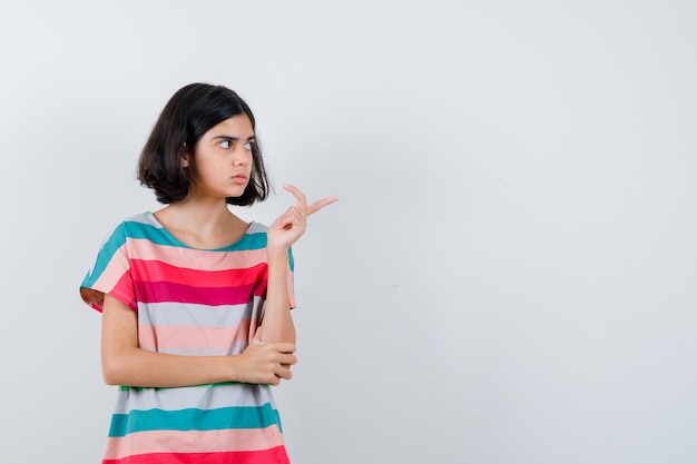 Little girl pointing right while holding hand on elbow in t-shirt, jeans and looking focused , front view.