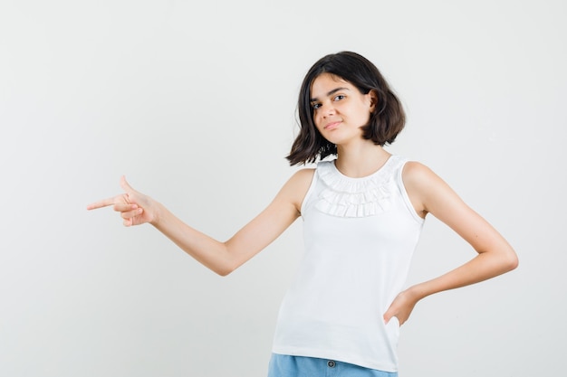 Little girl pointing to the side in white blouse, shorts and looking confident. front view.