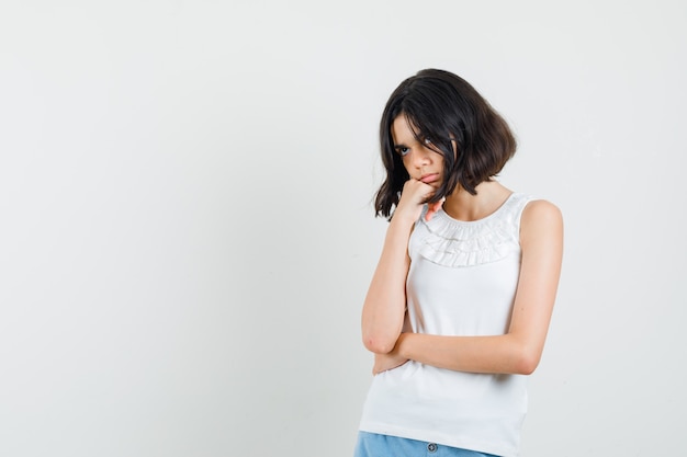 Little girl propping chin on raised fist in white blouse, shorts and looking pensive. front view.