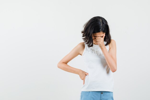 Little girl rubbing eyes and nose in white blouse, shorts and looking mournful , front view.