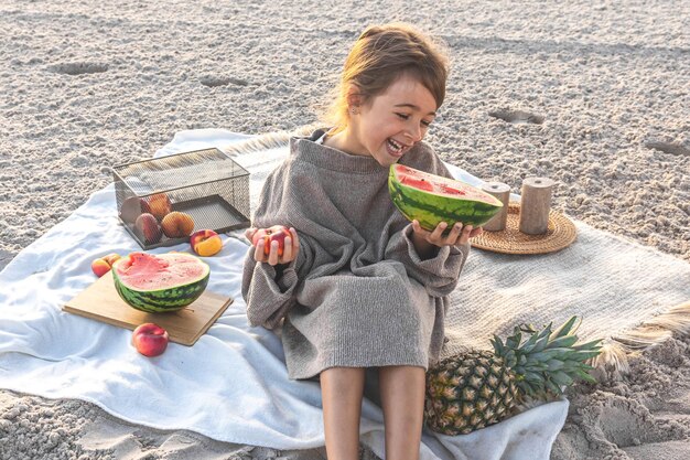 Little girl on the sandy seashore at a picnic with fruits