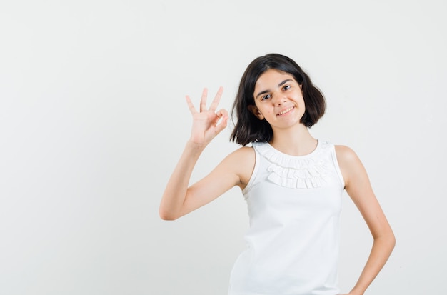 Little girl showing ok gesture in white blouse and looking satisfied , front view.