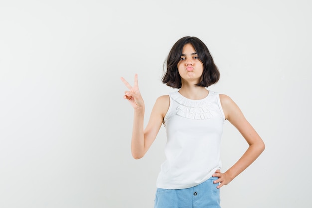 Little girl showing v-sign, pouting lips in white blouse, shorts and looking confident , front view.