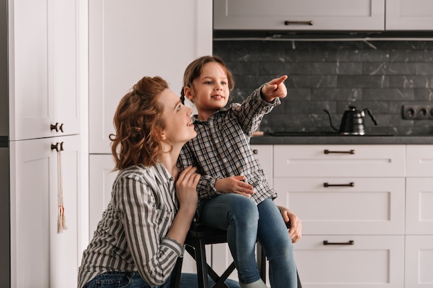 Little girl sits on chair and points her finger to side. Mom and daughter posing in kitchen.