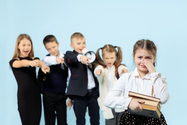 Little girl sitting alone on chair and suffering an act of bullying while children mocking. Sad young schoolgirl sitting on  against blue background.