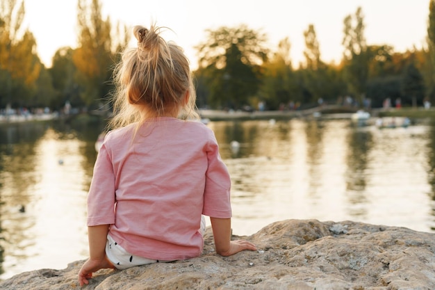 Little girl sitting on a huge stone in park near the lake