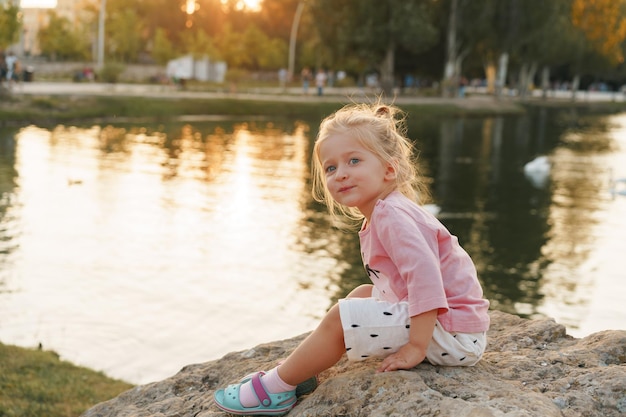 Little girl sitting on a huge stone in park near the lake