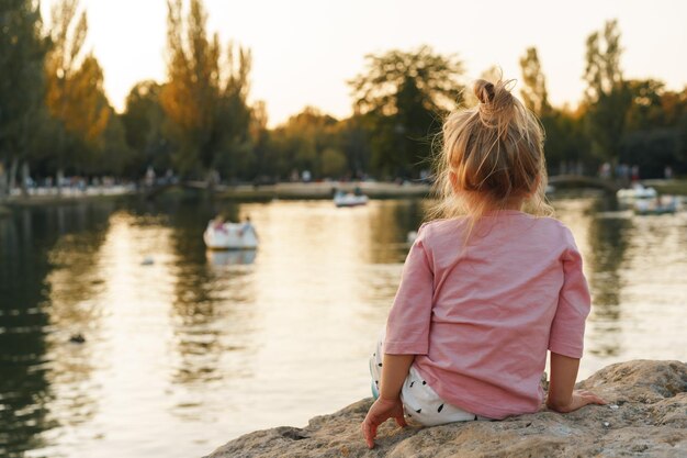 Little girl sitting on a huge stone in park near the lake