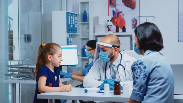 Little girl speaking with doctor during consultation for covid-19. Pediatrician specialist in medicine with protection mask providing health care services treatment examination in hospital cabinet