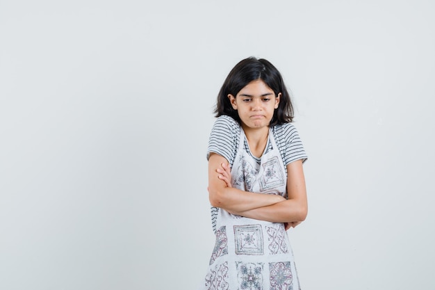 Little girl standing with crossed arms in t-shirt, apron and looking confused