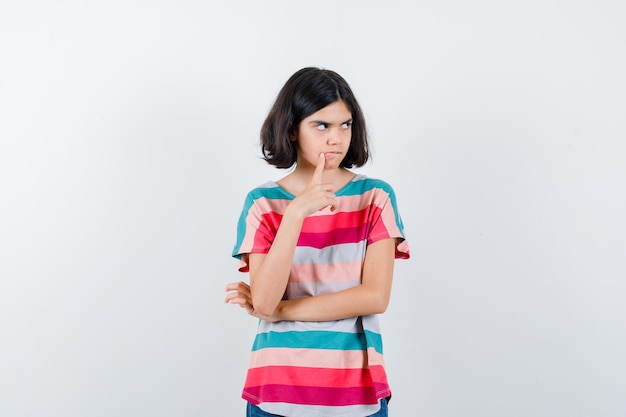 Little girl in t-shirt putting index finger near mouth, thinking about something and looking pensive , front view.