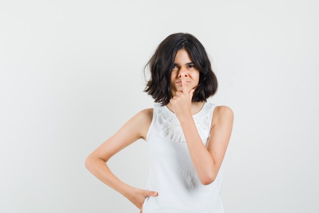 Little girl touching nose with finger in white blouse front view.