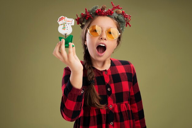 Little girl wearing christmas wreath in checked dress  holding christmas toy looking at camera happy and excited  standing over green background