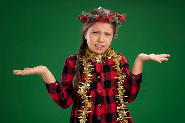 Free photo little girl wearing christmas wreath in checked dress  with tinsel around neck confused spreading arms to the sides