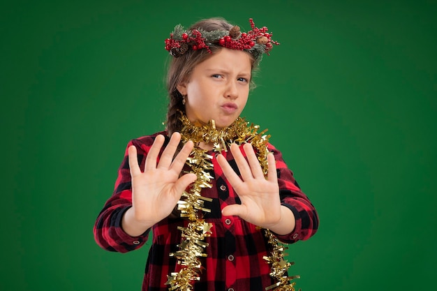 Free photo little girl wearing christmas wreath in checked dress  with tinsel around neck looking at camera worried making defense gesture with hands  standing over green background