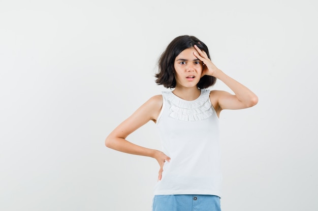 Little girl in white blouse, shorts holding hand to head and looking forgetful , front view.